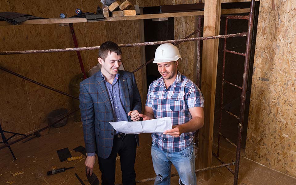 High Angle View of Young Male Architect and Construction Worker Foreman Inspecting Building Plans Together Inside Unfinished Building